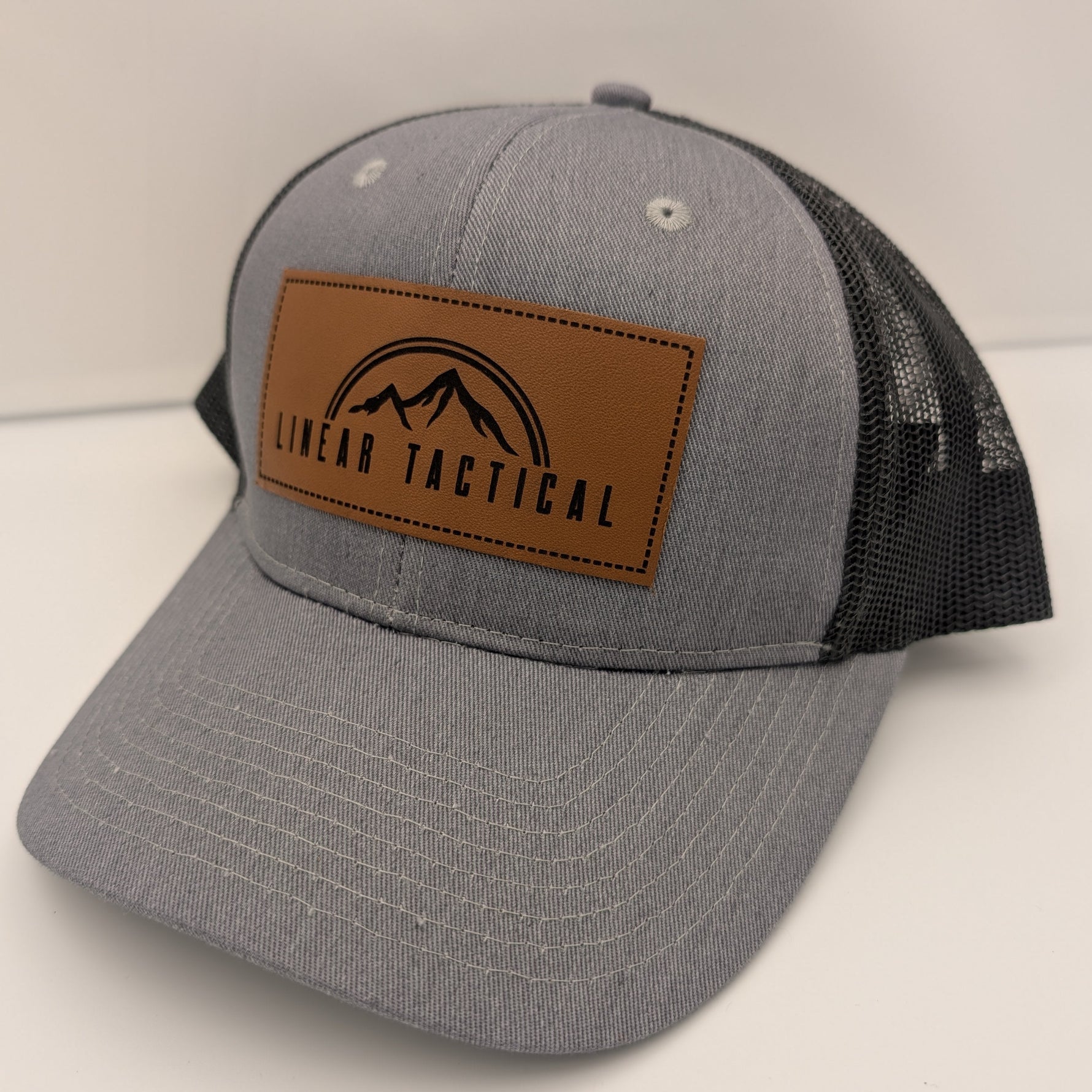 Gray trucker cap with a brown leather patch engraved with the Linear Tactical mountain peak logo on a white background